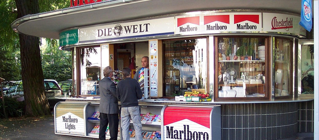 2 Personen stehen vor dem Bundesbüdchen in dem eine Person etwas anbietet